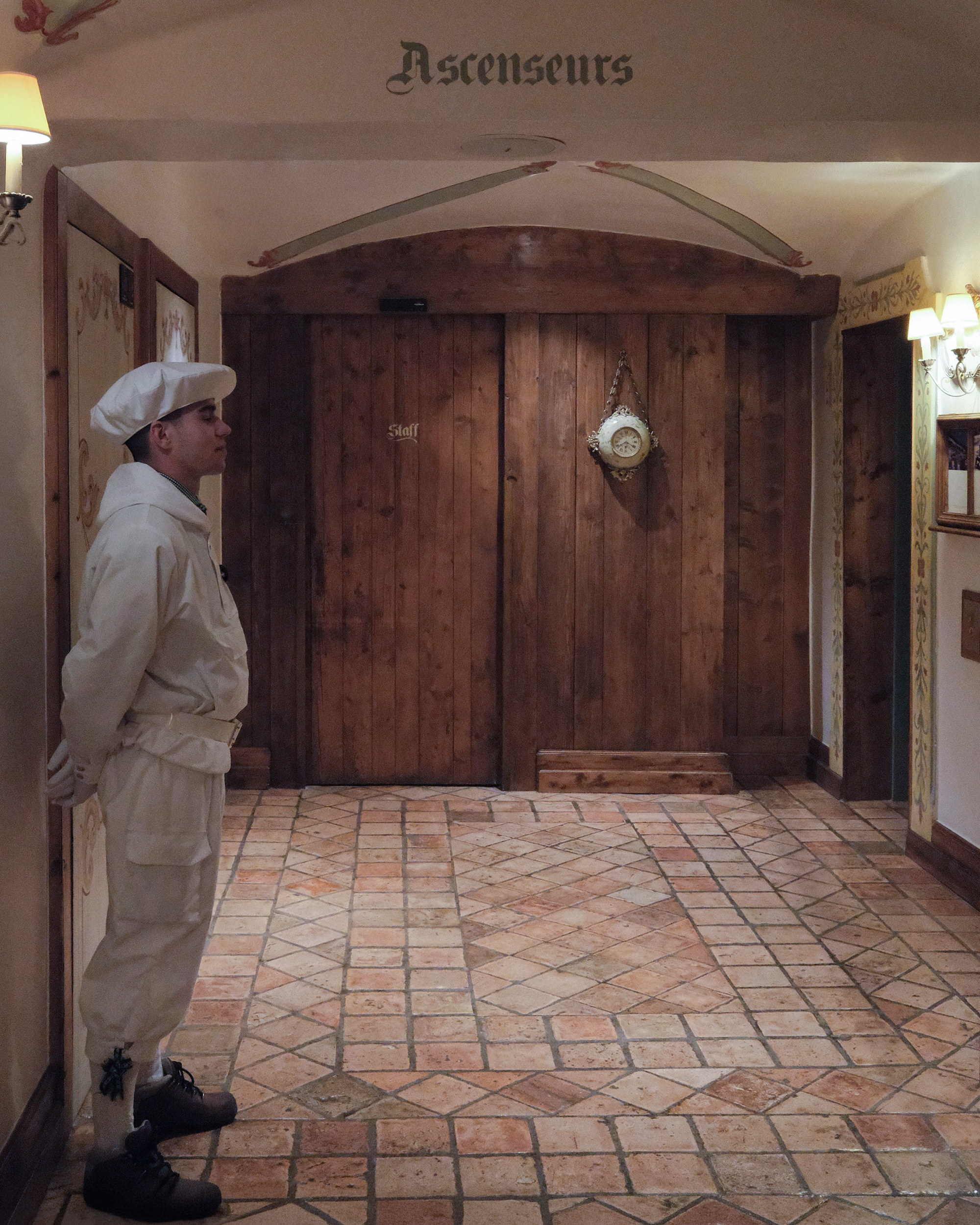A bellboy in white under the word Ascenseurs, a painted clock hung on the timber door