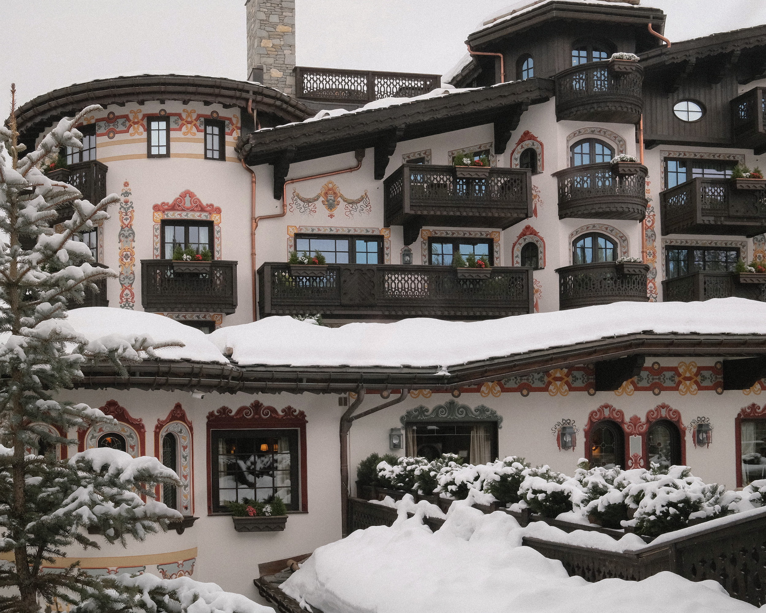 The elevation under fresh snow, painted frames and carved balconies rising to the stone chimney