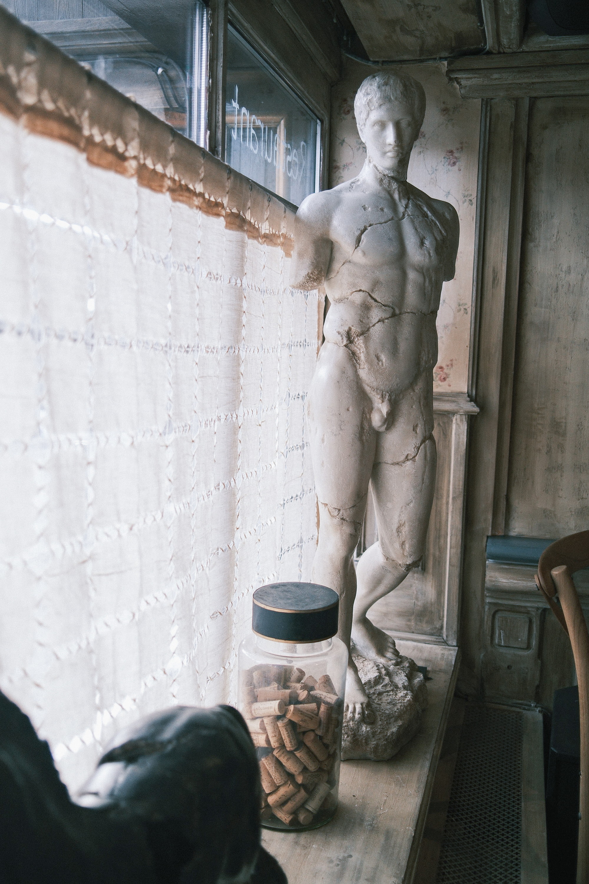 A plaster figure at the restaurant window, the lettering on the glass read from inside, a jar of corks at its feet