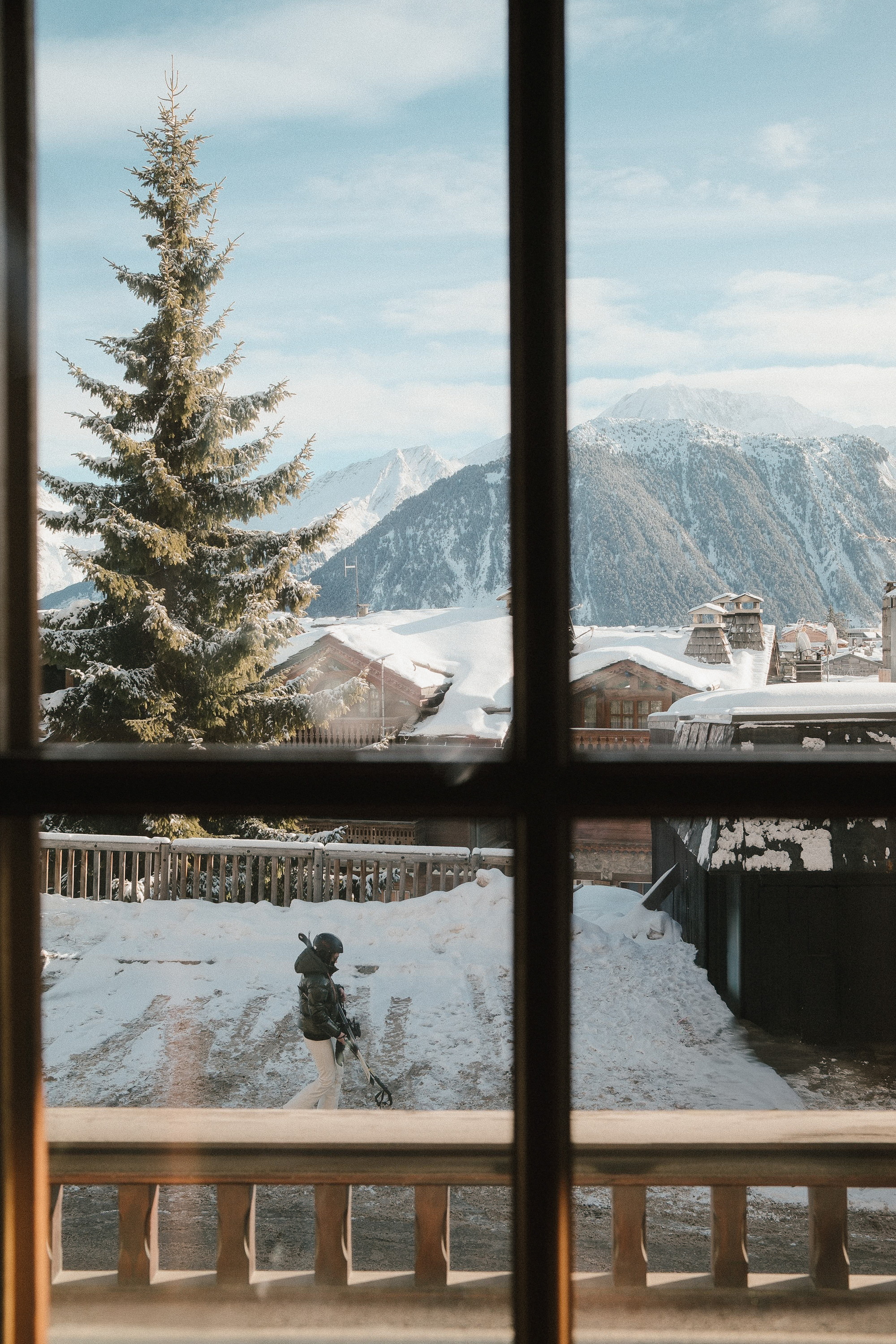 The village through the window, a skier crossing below, the peaks beyond the roofs
