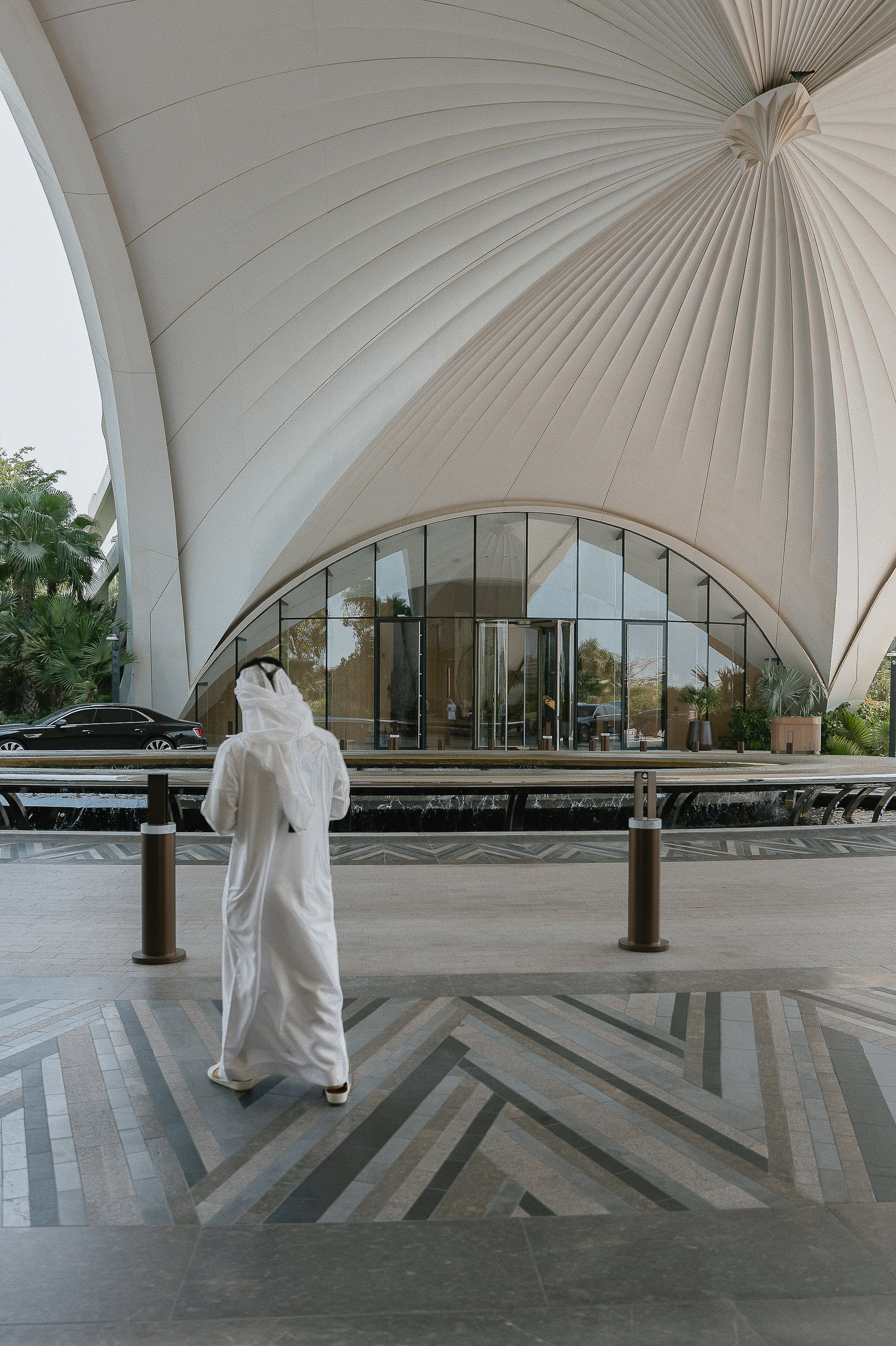 A figure in white at the porte-cochère, the canopy opening above the glass