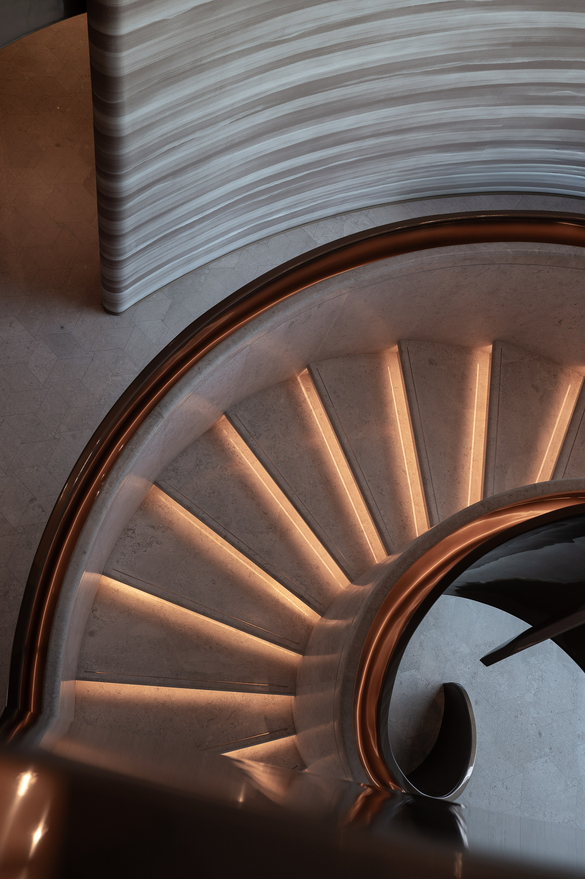 The stair from above, treads lit from beneath, the bronze rail turning