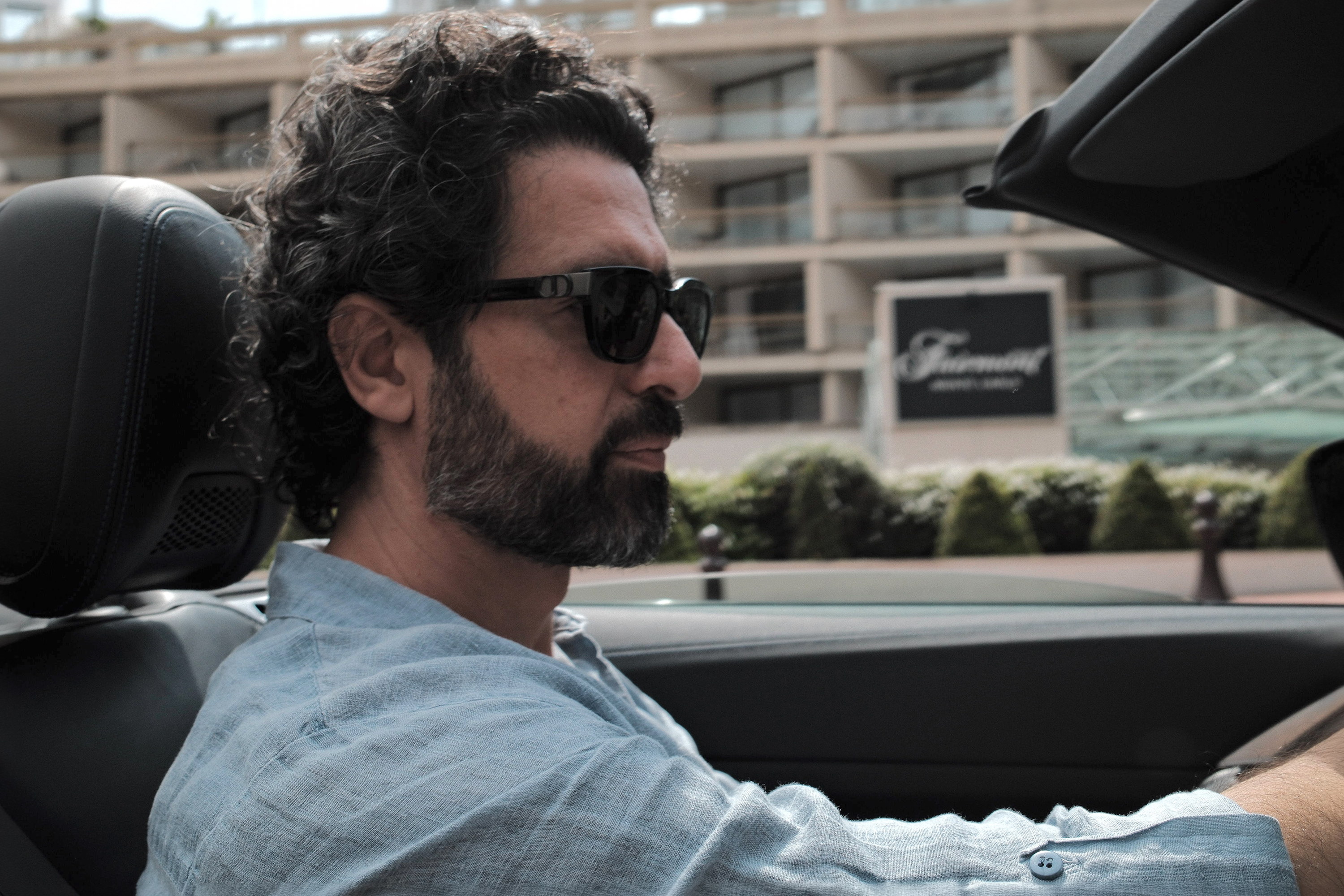 Linen and sunglasses at the wheel, the house falling away over his shoulder